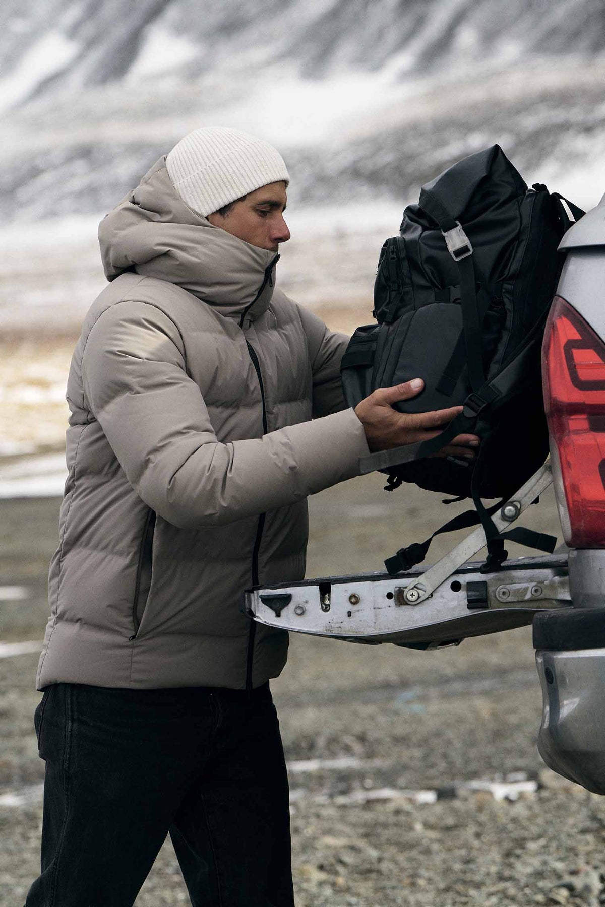 A man in a beige puffer jacket and white beanie loads a black backpack into the trunk of a silver vehicle in a snowy, mountainous outdoor setting.
