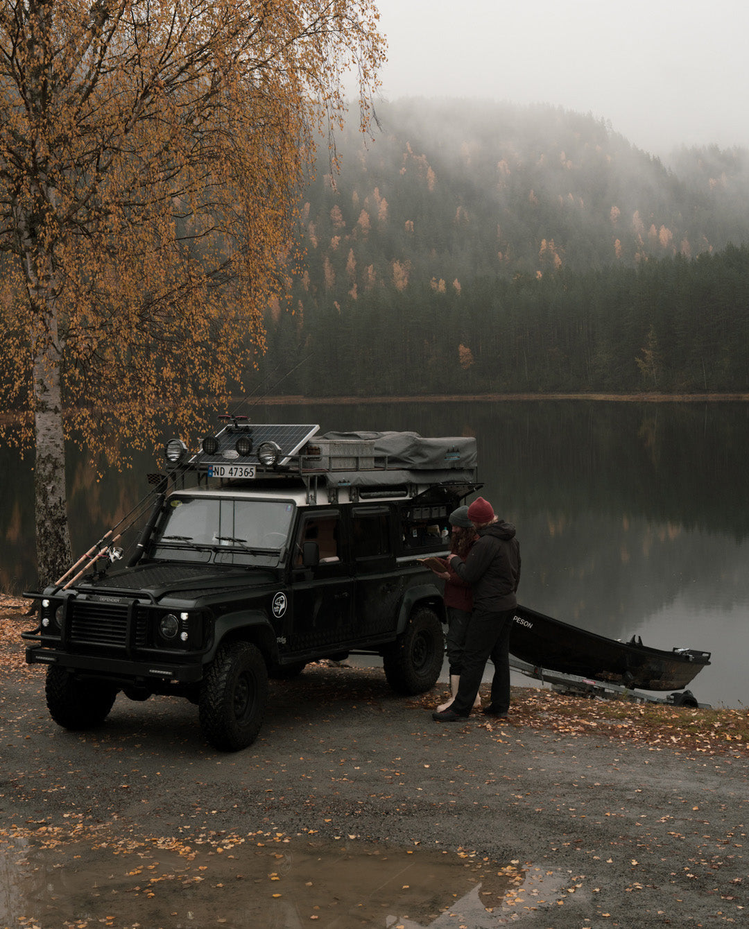 Land Rover Defender with a boat trailer parked by a misty lake under autumn birch trees