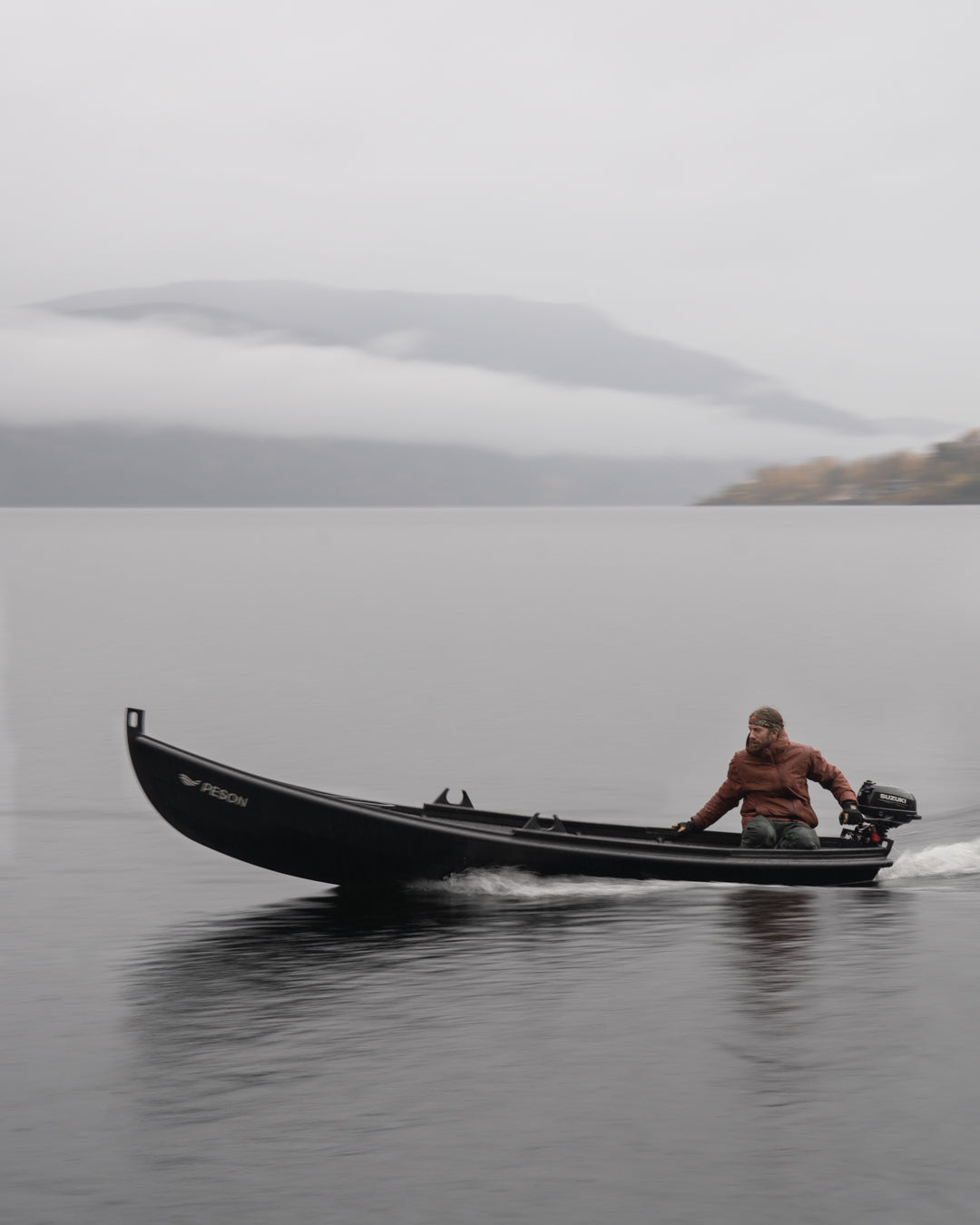 A long black boat with a small outboard crossing a still lake below low cloud