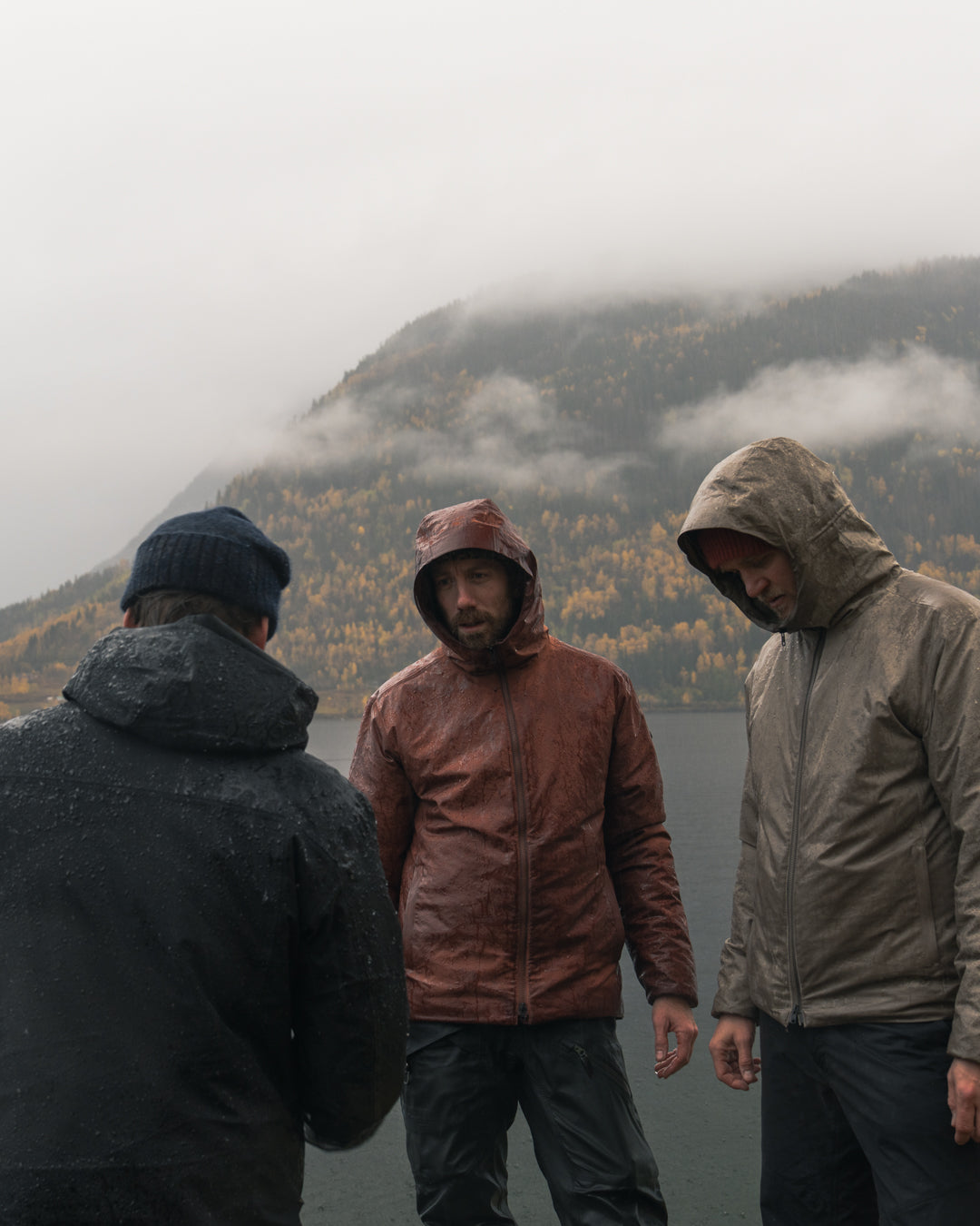 Three men in hooded jackets talking in the rain by the shore, with an autumn hillside in cloud behind them