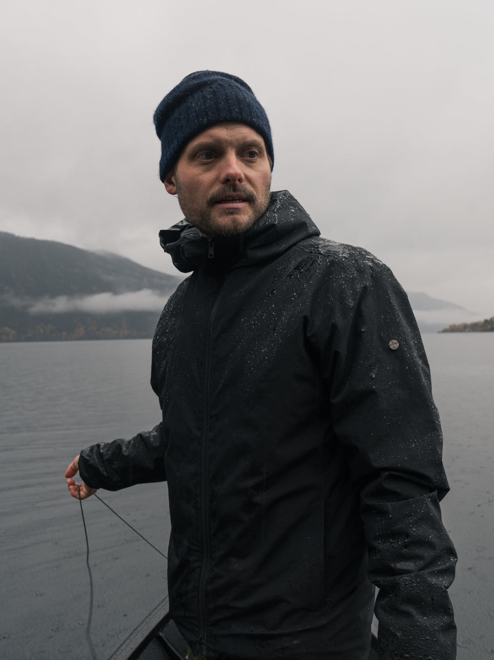 Man in a black jacket and navy beanie holding a line in the rain on the boat